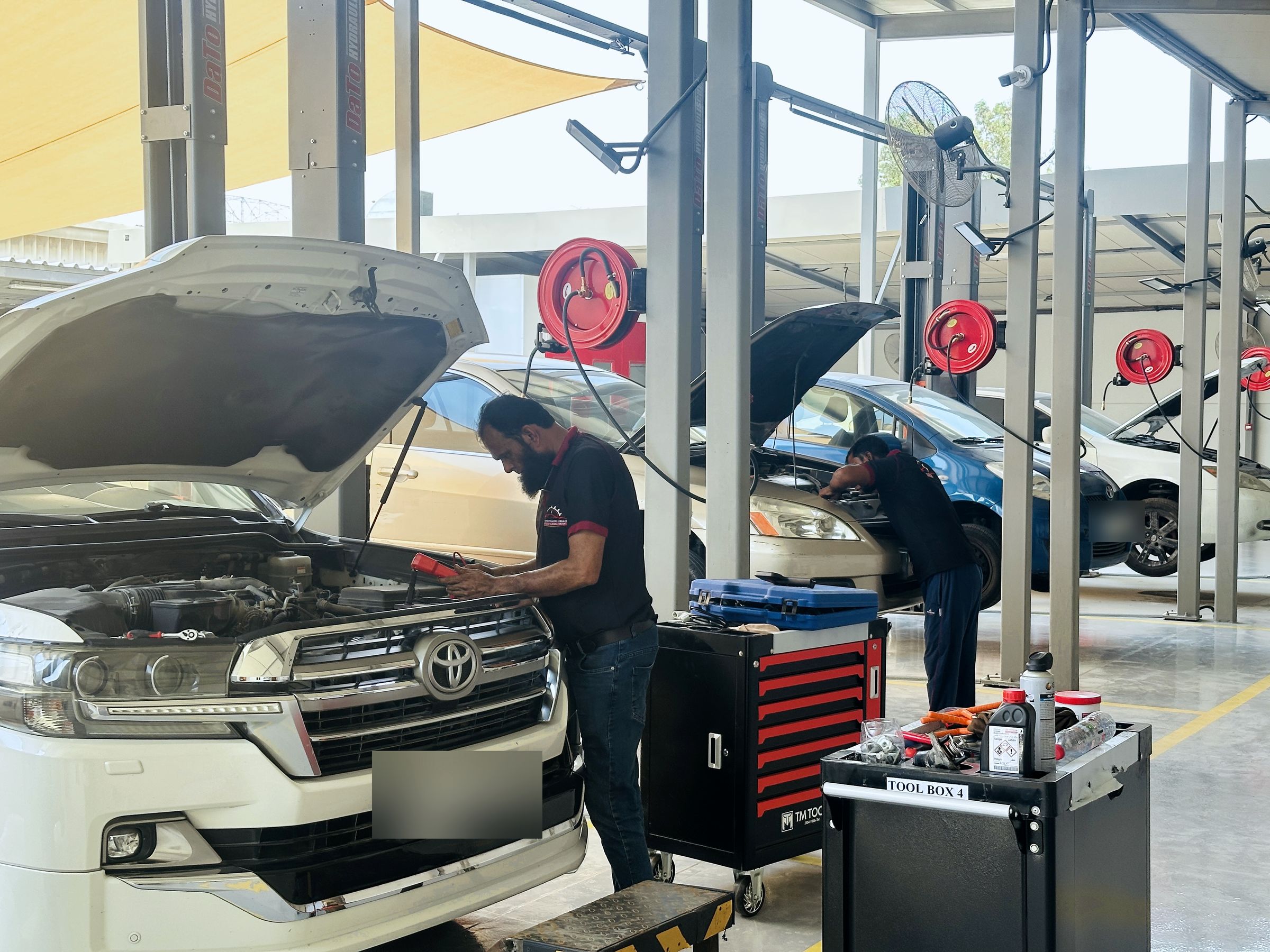 Two uniformed technicians at work: computer diagnostics on a Toyota Land Cruiser, red tool trolleys and hose reels in every bay